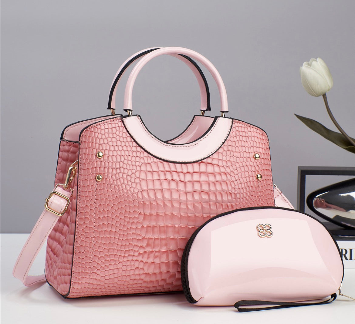 Pink crocodile-style textured handbag with rounded handles and shoulder strap, pictured beside a glossy pale pink pouch