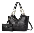Black crocodile-style handbag with metallic chain-link handles and shoulder strap, pictured beside a black envelope pouch